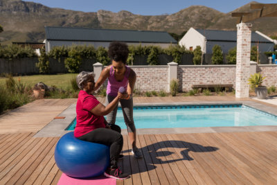 trainer training senior women in performing exercise on a swiss ball next to the swimming pool in the backyard of home